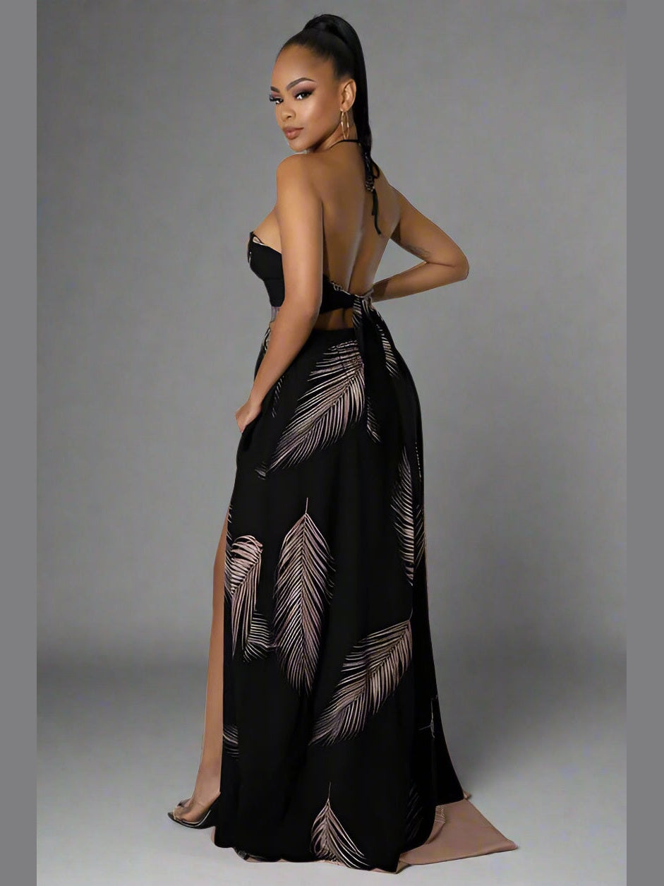 Black halter neck maxi dress with deep V-neckline, sleeveless cut, open back with a self-tie, elastic waistband, and a flowy, draped skirt. model standing to the side.